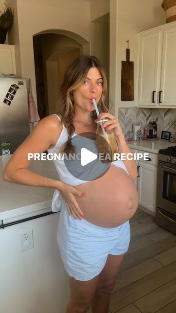 a pregnant woman holding a bottle in her right hand and brushing her teeth while standing in the kitchen