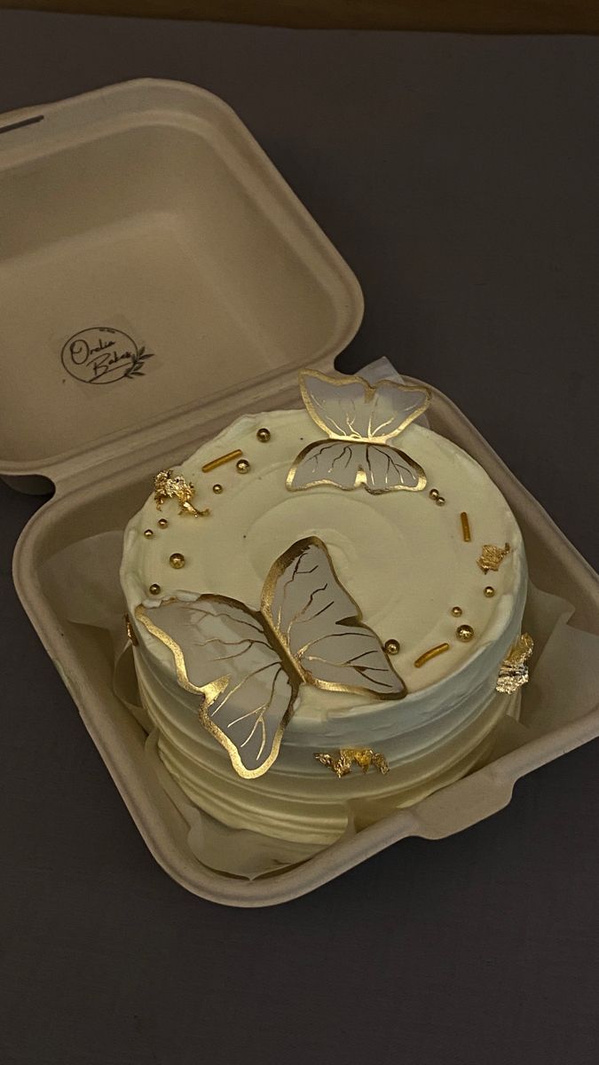 a cake in a plastic container with gold butterflies on the top and sides, sitting on a table