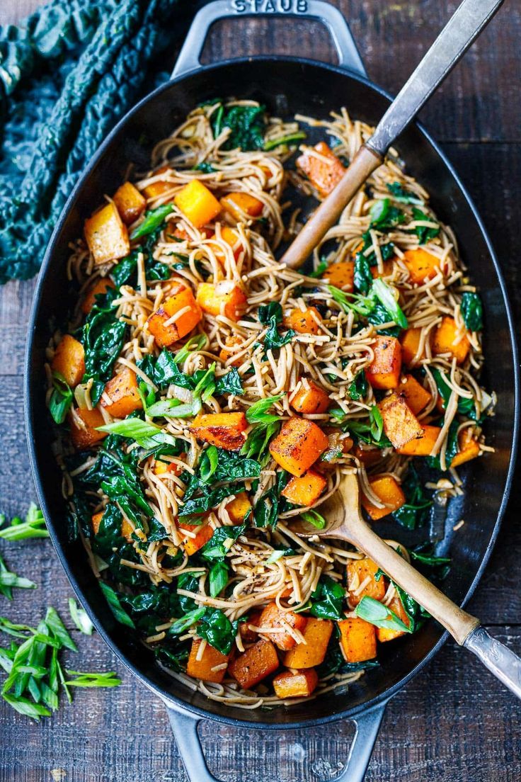 a skillet filled with noodles, carrots and spinach on top of a wooden table