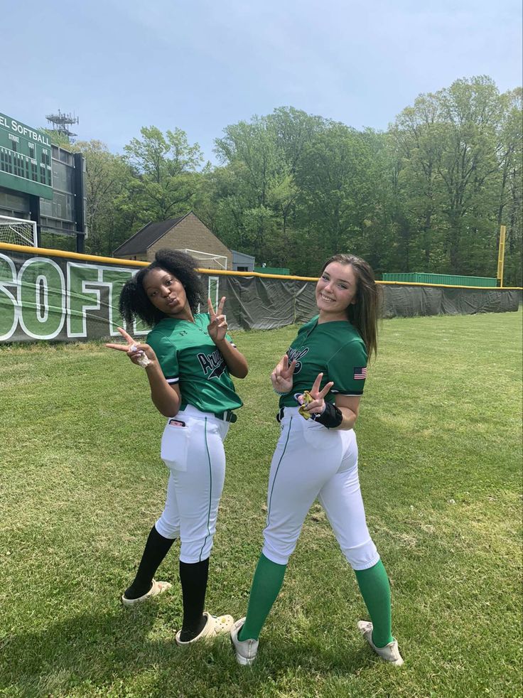 two girls in green and white baseball uniforms standing on the field with their hands up