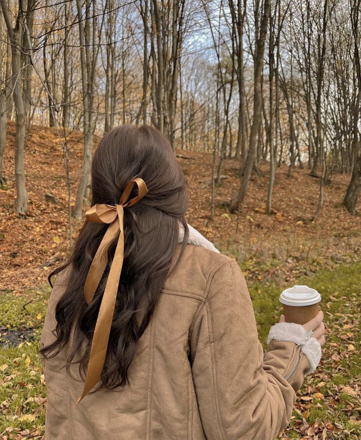 a woman walking in the woods holding a cup of coffee and wearing a brown bow around her neck