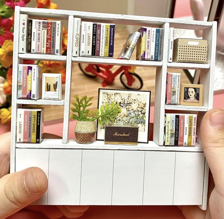 a person holding up a miniature book shelf with books and pictures on it's sides