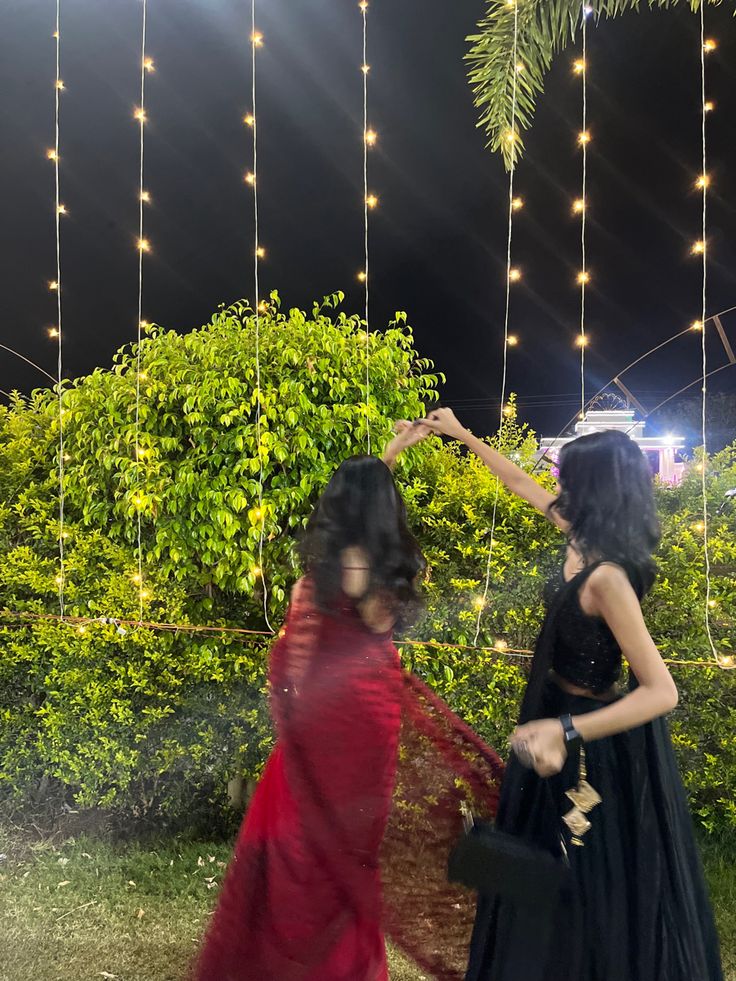 two women dressed in black and red dancing outside at night with string lights strung above them