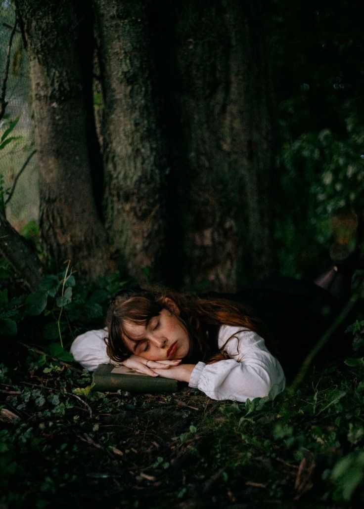 a woman laying on the ground next to a tree with her eyes closed and head down