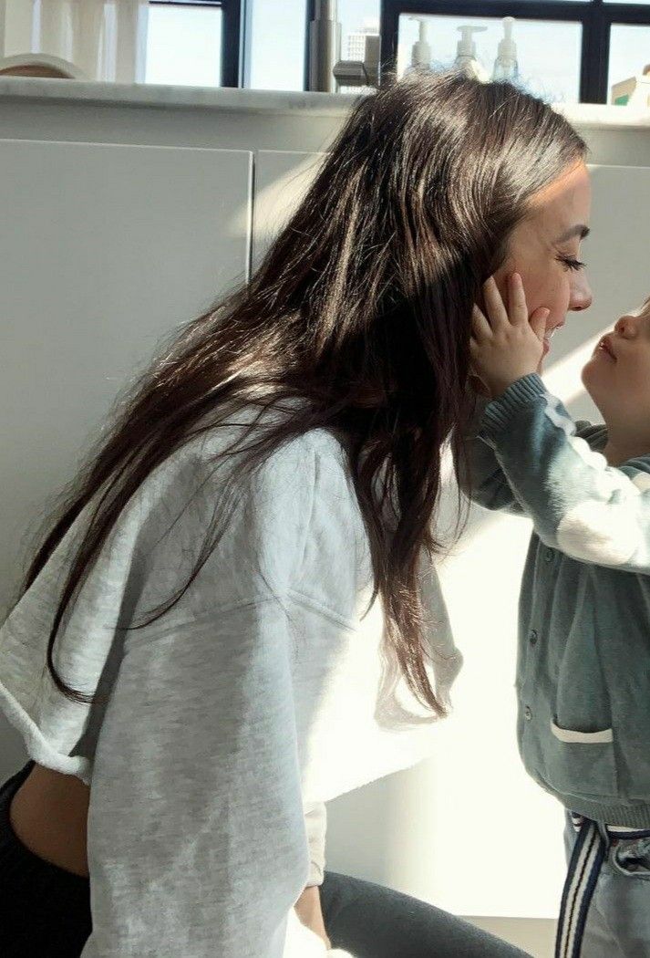 a woman talking on a cell phone next to a little boy with long hair in the kitchen