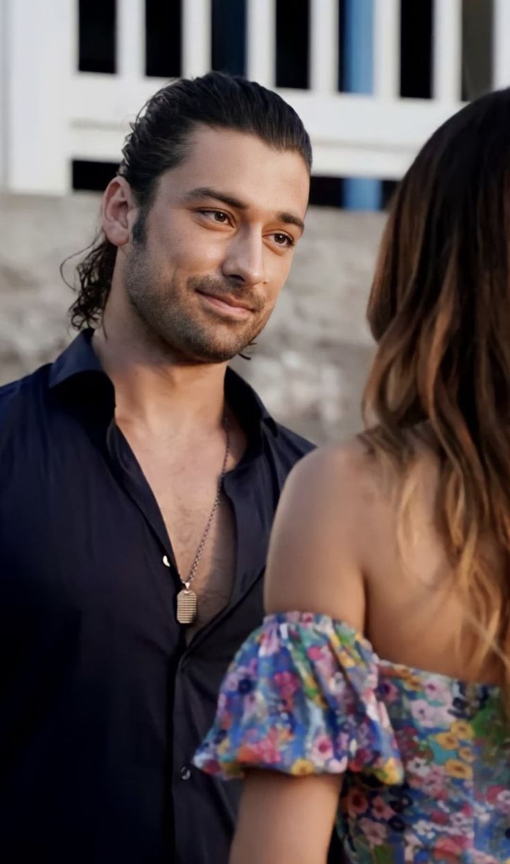 a man with long hair standing next to a woman in a floral dress and looking at each other