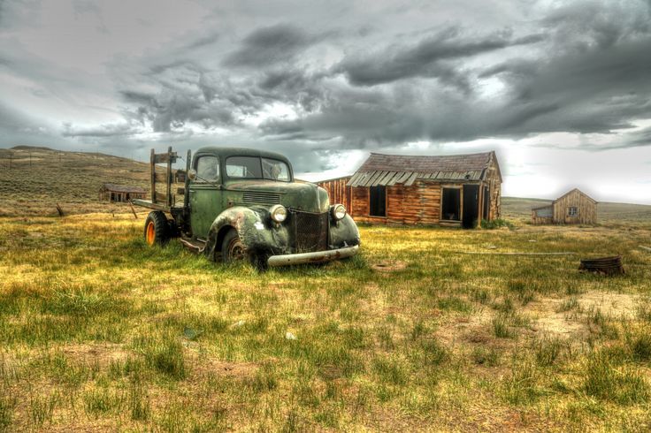 an old truck is parked in the middle of a grassy field next to two buildings