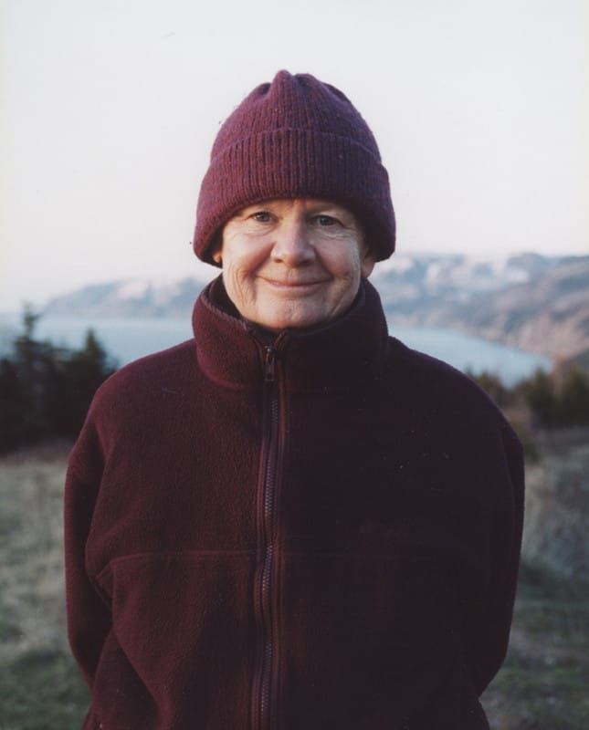 an older woman wearing a maroon jacket and hat standing in front of a lake with mountains in the background