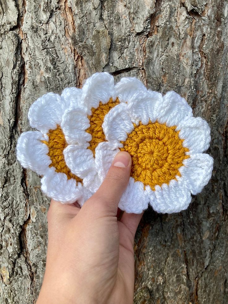 a hand holding two crocheted white and yellow flowers on top of a tree