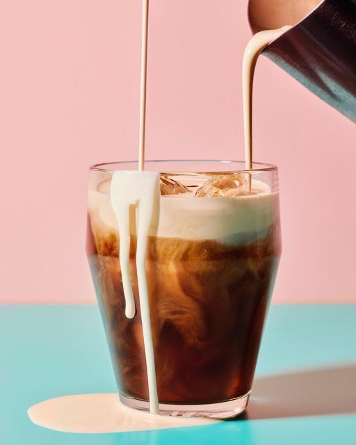 a person pouring milk into a glass filled with ice cream and iced coffee on top of a blue table