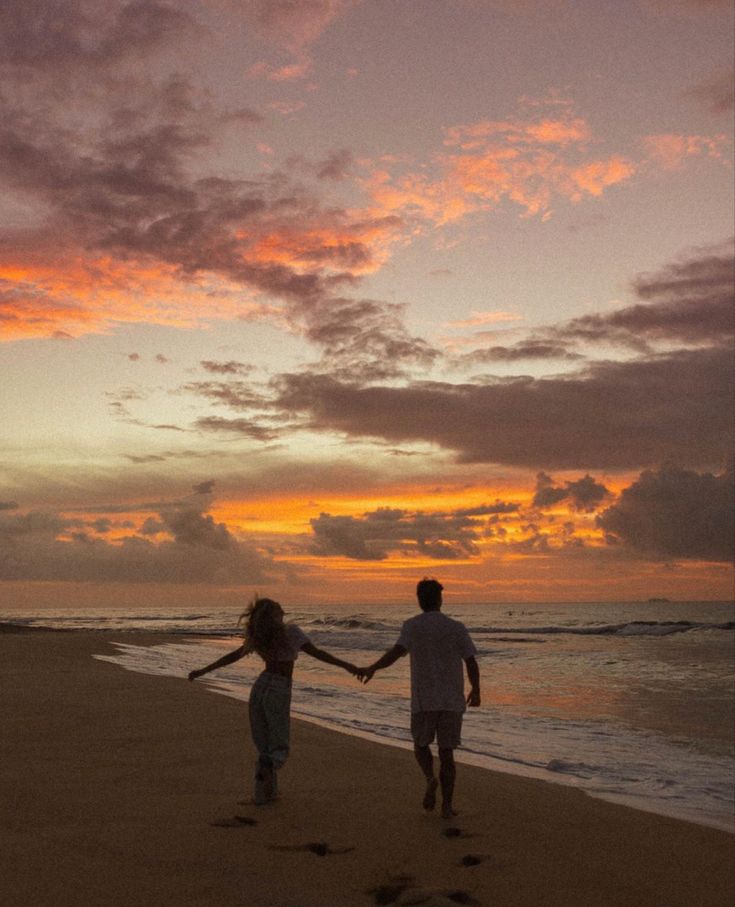 two people holding hands walking on the beach at sunset with footprints in the sand as the sun sets