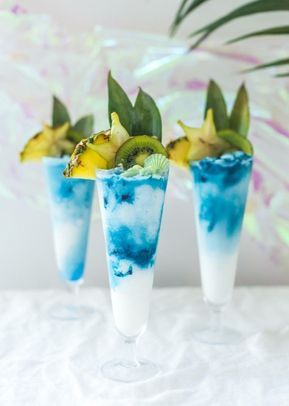 three glasses filled with blue liquid and fruit on top of a white table next to a palm tree