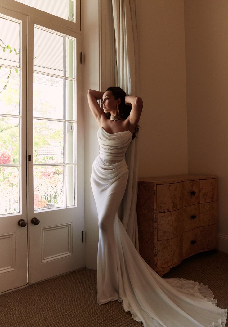 a woman in a white dress leaning against a window with her arms behind her head