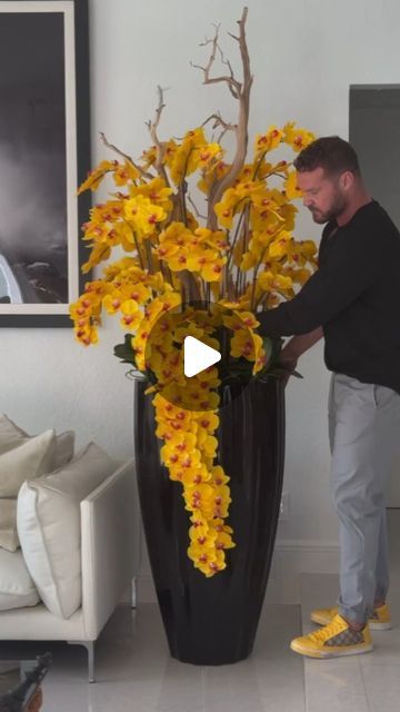 a man standing next to a large vase filled with yellow flowers on top of a white floor