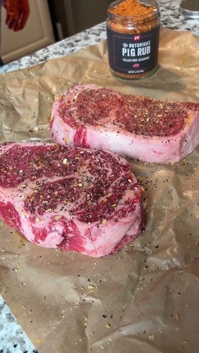 two raw steaks sitting on top of a piece of wax paper next to a jar of seasoning