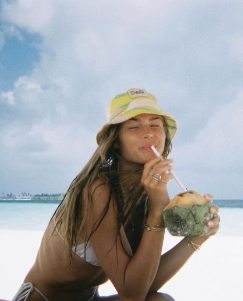 a woman sitting on the beach with a coconut drink in her hand and drinking from a straw