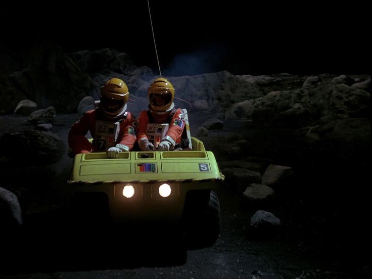 two people in the back of a green vehicle with lights on and one person wearing safety gear