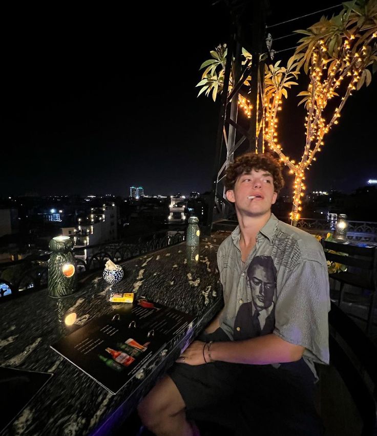 a young man sitting on top of a wooden table next to a tree filled with lights