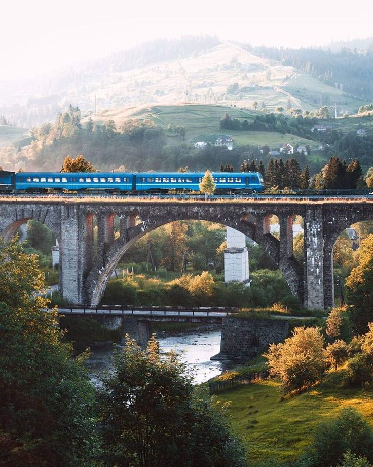 a blue train traveling across a bridge over a river next to lush green hillside covered with trees