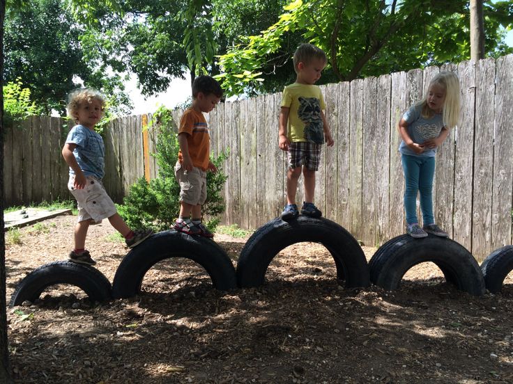 four children standing on top of old tires in the dirt near a fence and tree