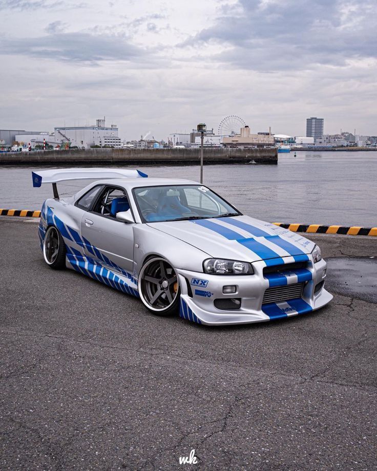 a silver car with blue stripes parked in front of a body of water on a cloudy day