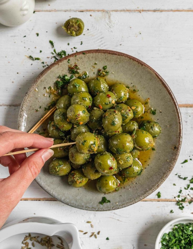 someone holding chopsticks over a bowl filled with green vegetables on a white table