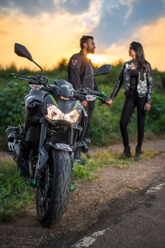 a man and woman standing next to a motorcycle on a dirt road with the sun setting in the background