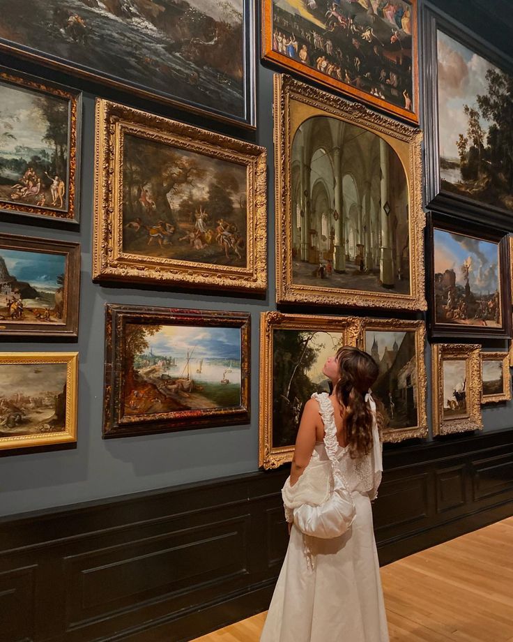 a woman in a white dress looking at paintings on the wall and wooden flooring