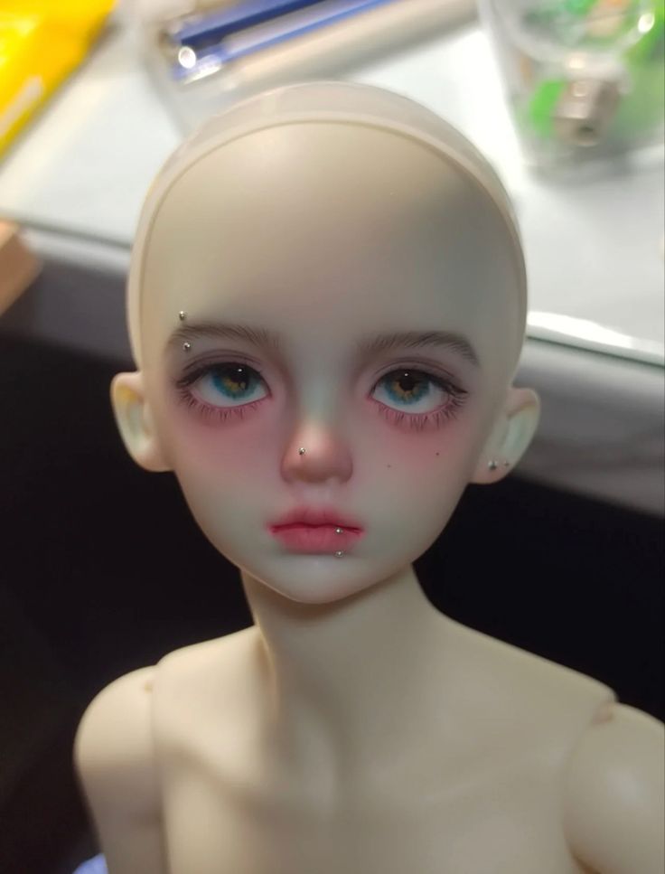 a white doll with blue eyes and piercings on it's head sitting in front of a desk