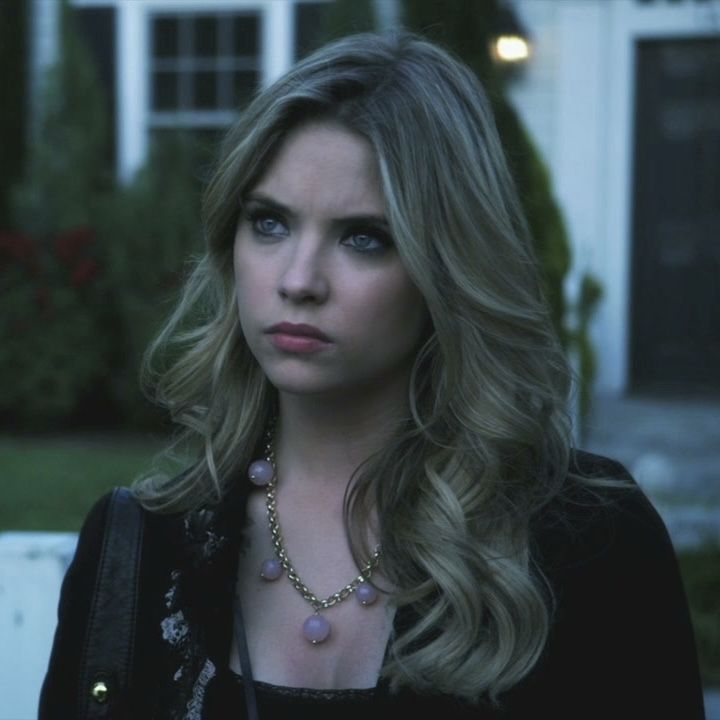 a woman with long blonde hair wearing a black jacket and necklace standing in front of a house