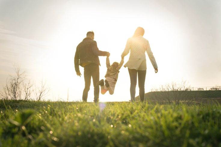 two adults and a child holding hands while standing in the grass on a sunny day