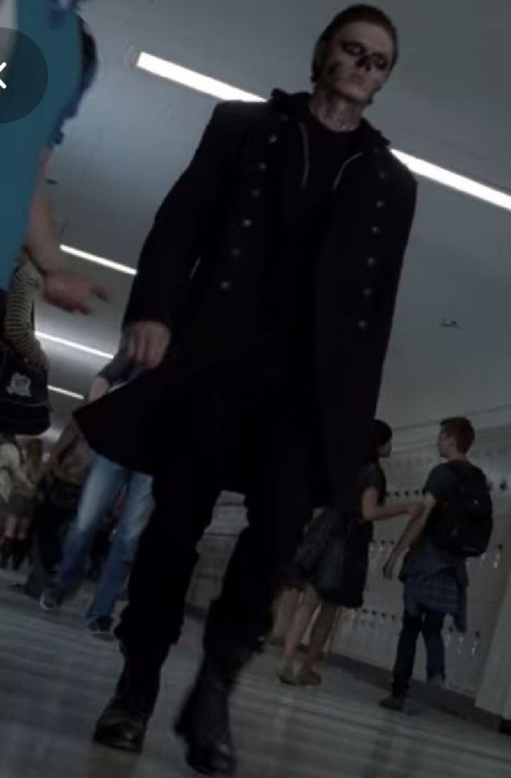 a man in a black coat and sunglasses walking down a hall with other people behind him