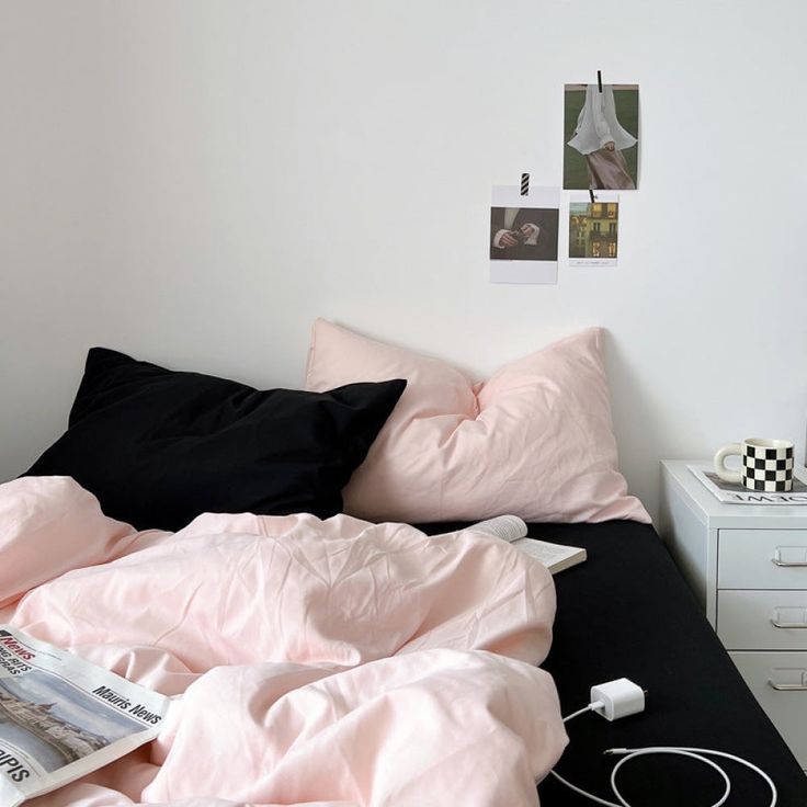 an unmade bed with pink sheets and black comforter in a white walled room