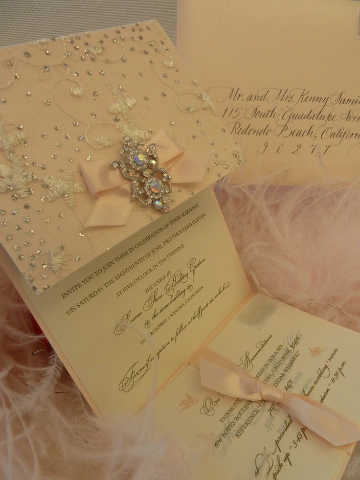 the wedding card is laying on top of some feathers and pink flowers, with an embellished brooch