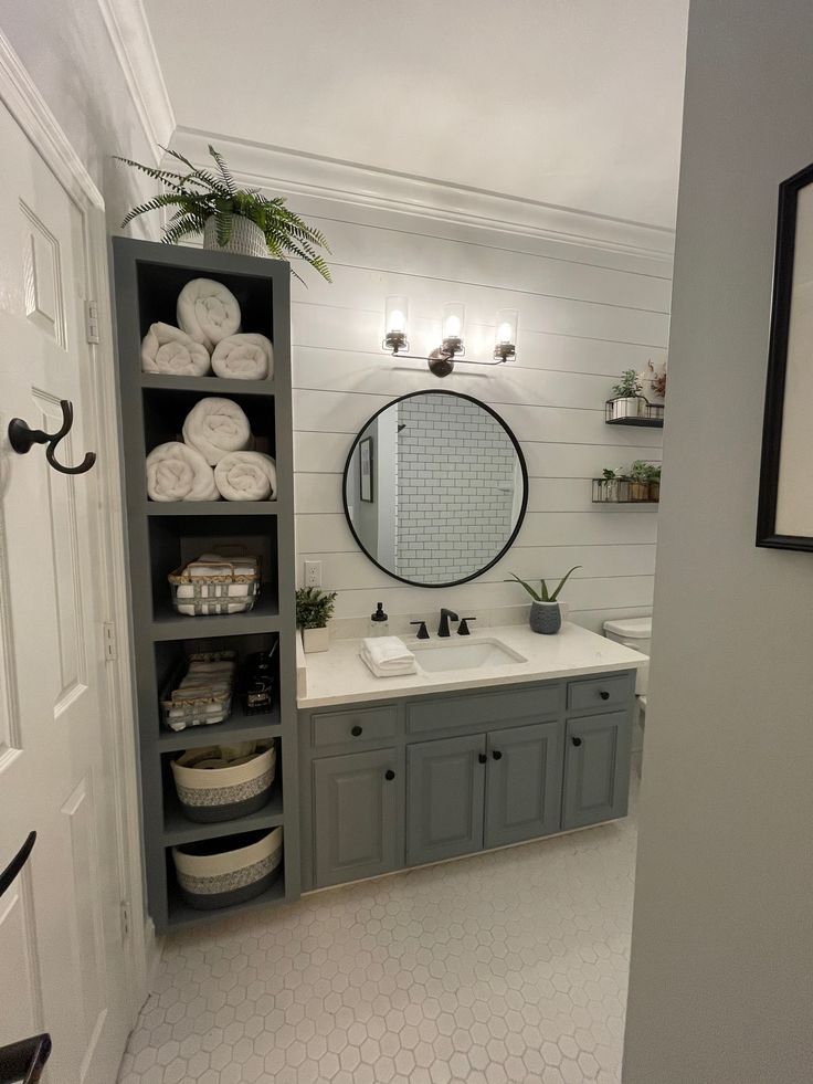 a bathroom with gray cabinets and white towels on the shelves, along with a large round mirror