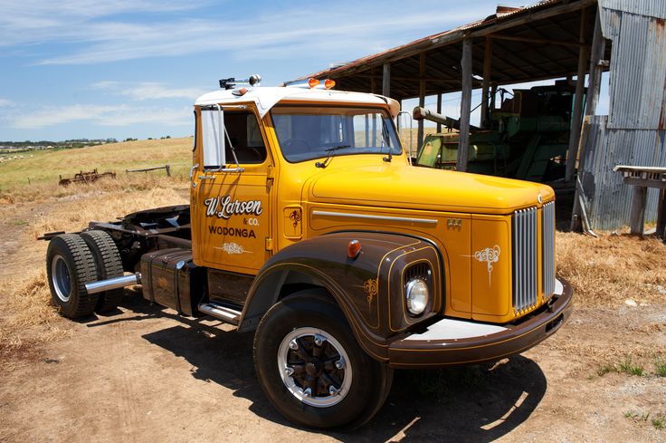 an old yellow truck is parked in front of a barn on the side of a dirt road