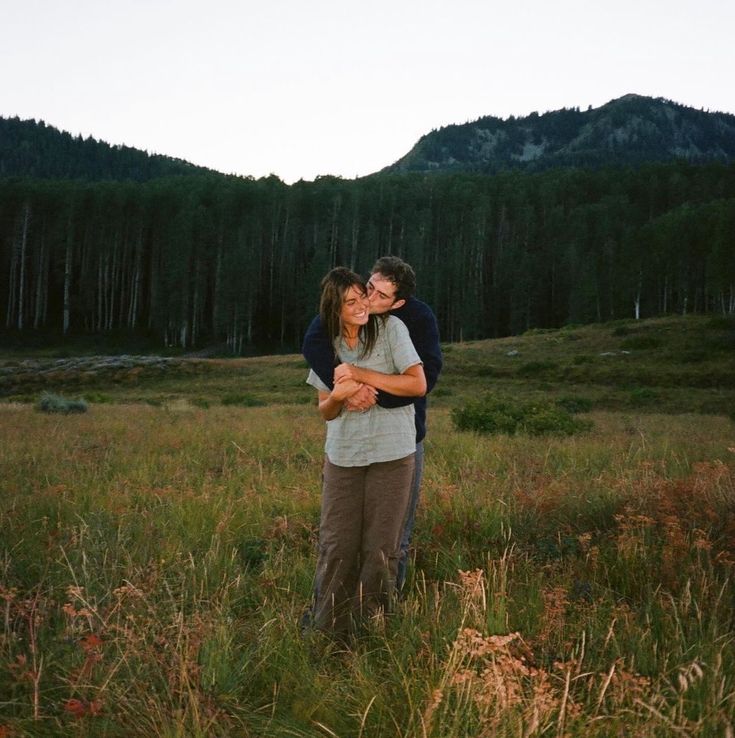 a man and woman hugging in the middle of a field with mountains in the background