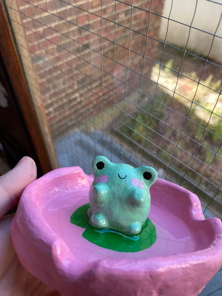 a small toy frog sitting in a pink bowl with green water on it's side