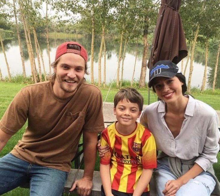 a man, woman and boy sitting on a bench in front of a lake with trees