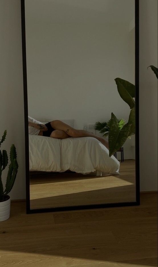 a woman laying on a bed in front of a large mirror with plants behind her