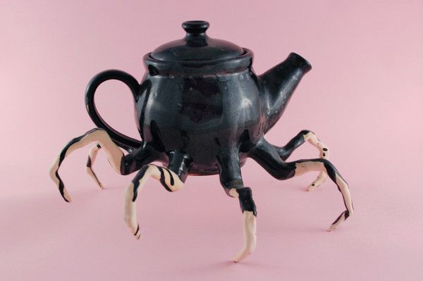 a black teapot with white legs and claws on it's side, sitting in front of a pink background