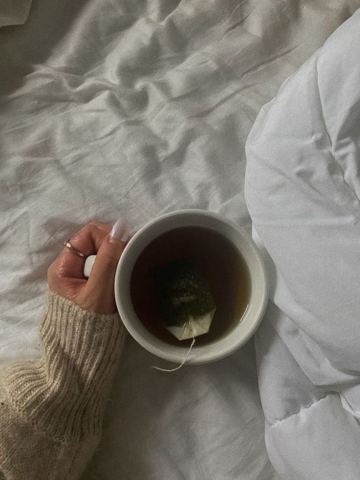 a person holding a cup of tea on top of a white bed covered in sheets