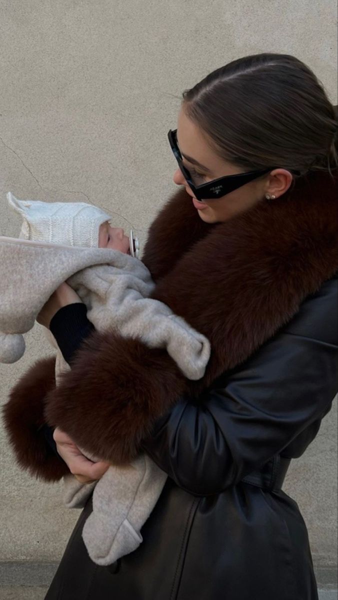 a woman holding a baby in her arms while she is wearing a black coat and sunglasses