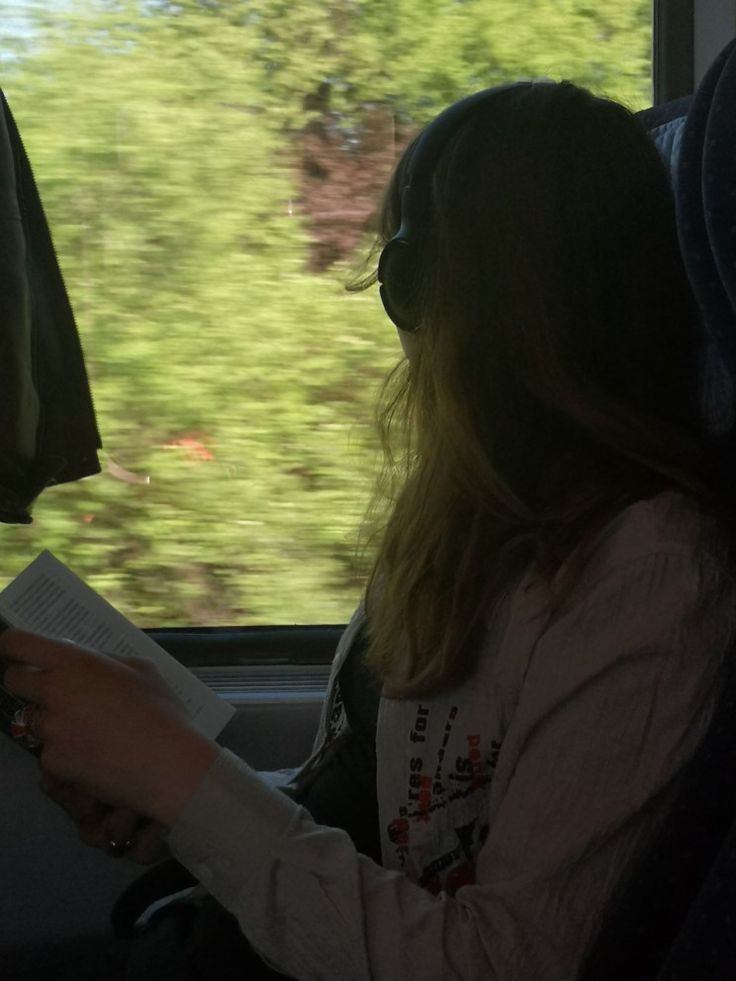 a woman sitting on a bus reading a book and looking out the window at trees