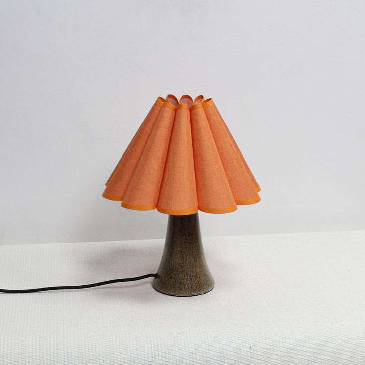 an orange lamp shade sitting on top of a white cloth covered table next to a black cord