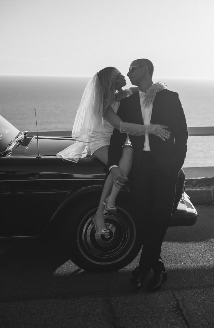 a bride and groom kissing in front of an old car by the ocean on their wedding day