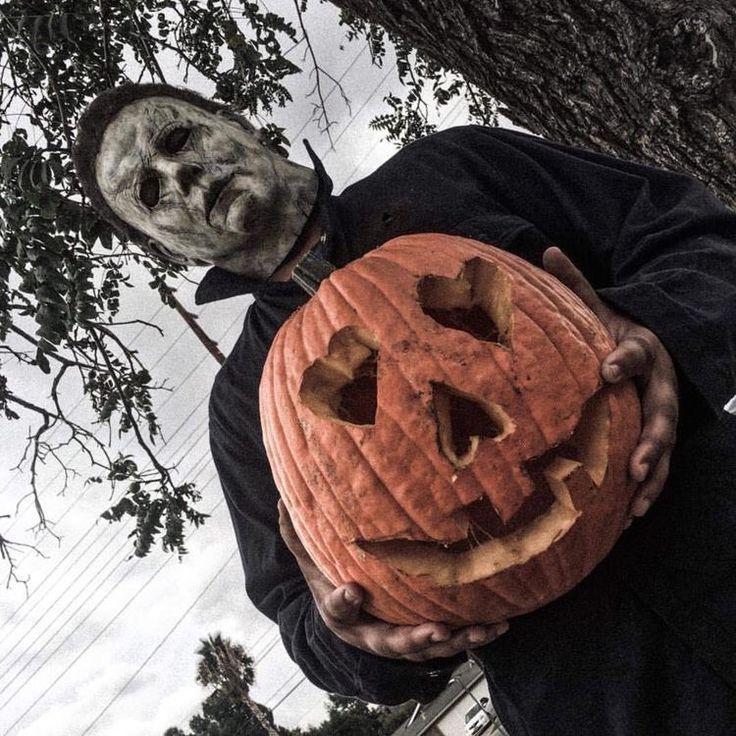 a man holding a carved pumpkin in front of a tree with a creepy face on it