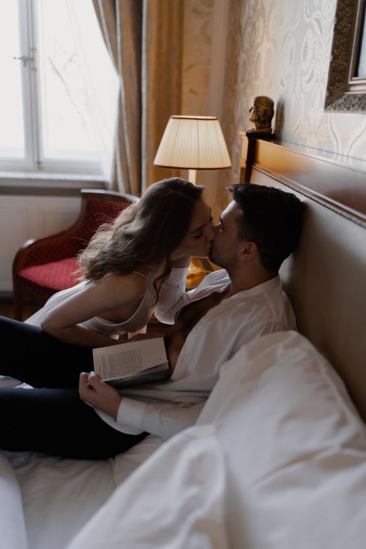 a man and woman laying in bed kissing while holding an open book with their hands