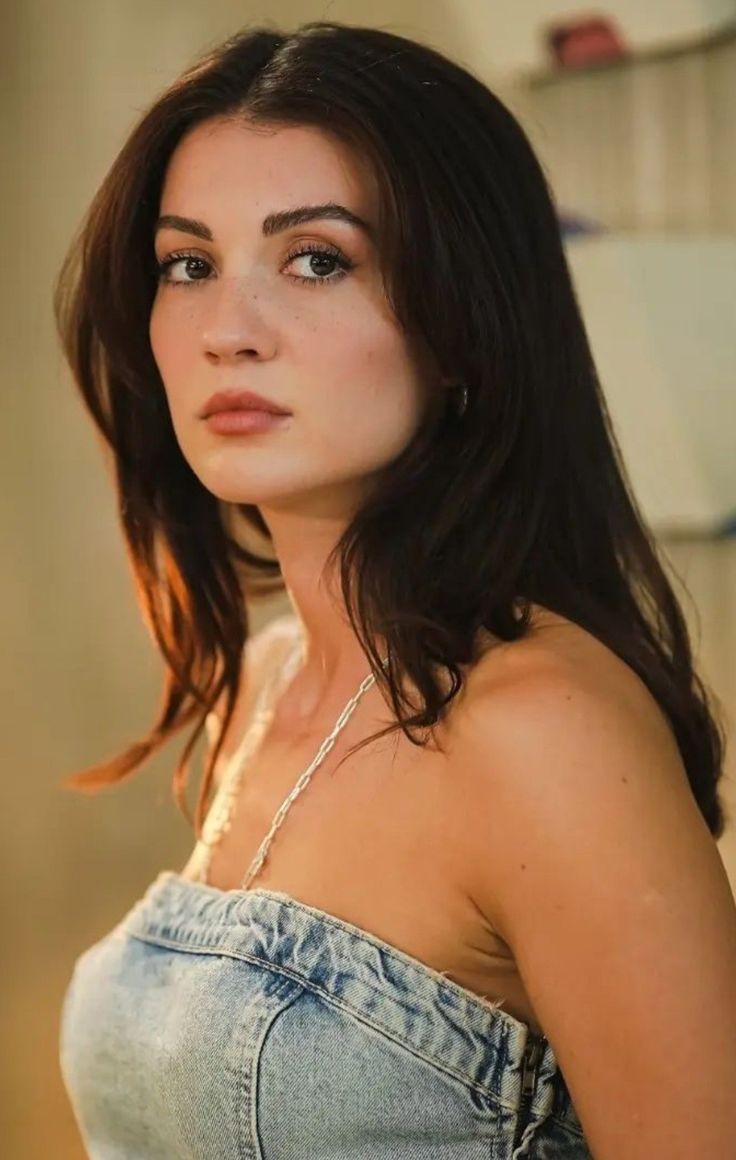 a woman with long dark hair wearing a denim top and jeans is looking at the camera