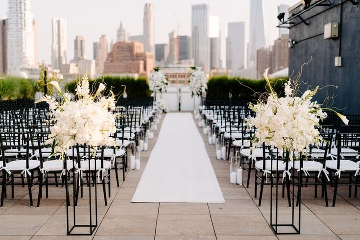 an outdoor ceremony setup with white flowers and black chairs on the ground in front of a city skyline